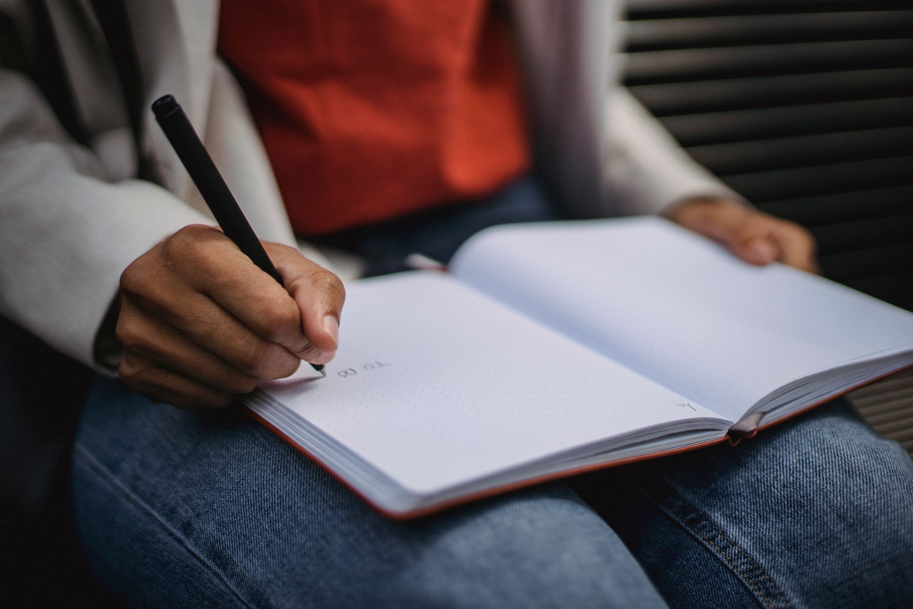 A person writing by hand in a notebook at a desk near a window